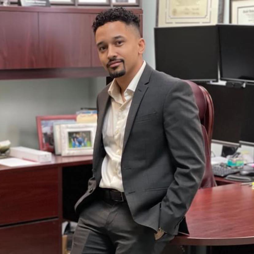 A man in a suit sits in an office chair behind a desk.